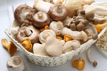 Different raw mushrooms in wicker basket on white tiled table, closeup