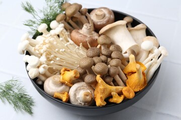 Different raw mushrooms in bowl and dill on white tiled table, closeup