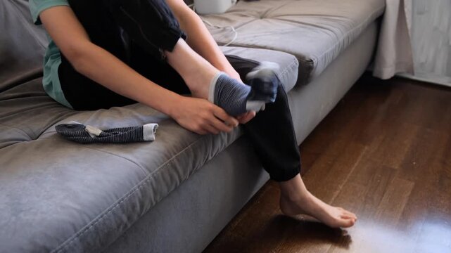 Side view of a teen boy sitting on a sofa and putting on striped socks as part of a morning routine at home.