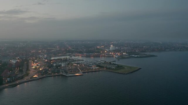 Aerial night view of Thisted, Denmark. Town lights, marina, Limfjord coastline, and surrounding neighborhoods create a tranquil Nordic cityscape under a twilight sky
