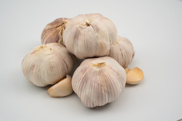 Five garlic bulbs are arranged together on a plain white surface, highlighting their papery skin and unique shape. A few garlic cloves are scattered nearby, emphasizing their freshness.