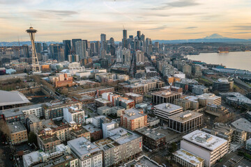 Aerial panoramic view of Seattle waterfront with city skyline, Mount Rainier in the background, Elliott Bay, piers, ferries, modern skyscrapers, trains, and dramatic sunset sky over Puget Sound