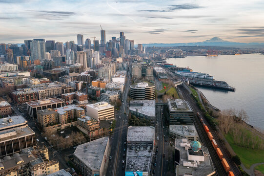 Aerial panoramic view of Seattle waterfront with city skyline, Mount Rainier in the background, Elliott Bay, piers, ferries, modern skyscrapers, trains, and dramatic sunset sky over Puget Sound