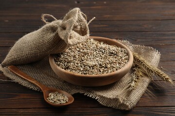 Rye grains in bowl, spoon, spikes and burlap cloth on wooden table, closeup