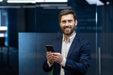 Portrait of a smiling young businessman in a suit standing in an office, holding a phone, looking at the camera