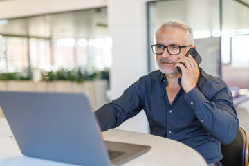 Smiling mature businessman holding smartphone sitting in office. Middle aged manager ceo using cell phone mobile apps and laptop.