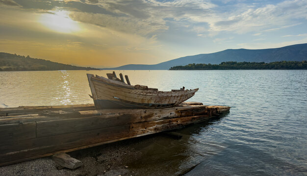 Weathered wooden fishing boat resting on pier at coastline during golden sunset with calm sea and distant mountain backdrop