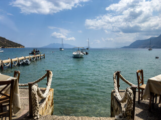 Seaside taverna wooden chairs and tables overlooking anchored boats within the clear water of the Saronic Gulf, Greece