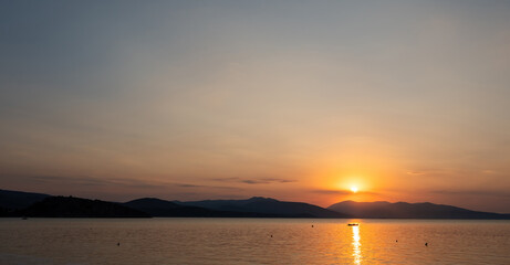 Tranquil fishing boat at sunset over Tolo Bay Greece. Glowing orange sky and distant mountain silhouettes reflecting on calm sea water