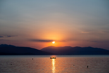 Tranquil fishing boat at sunset over Tolo Bay Greece. Glowing orange sky and distant mountain silhouettes reflecting on calm sea water
