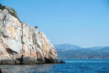 Fototapeta premium A rugged rocky coastal cliff rises above the calm blue sea near Tolo, blue sky