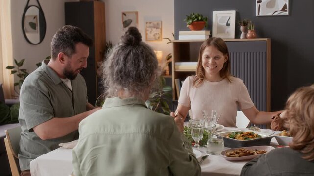 Happy extended family consisting of married couple, senior woman and two children filling plates with food after sitting down at table for holiday dinner