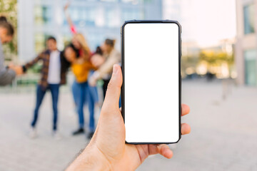 Hand holding a smartphone with blank white screen, perfect for mockup, with a group of friends celebrating in the background