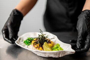 Leaf-shaped plate with rice, seaweed, vegetables, and lime wedge held by gloved hands. Minimalist background. Hygienic food presentation in professional kitchen setting