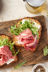 Grilled toasts with ham, cheese and arugula on wooden cutting board on light textured background top view. Delicious breakfast or snack