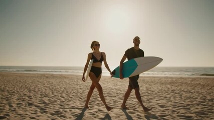 Cheerful young couple carrying surfboards while walking along sandy beach at golden hour sunset, enjoying summer vacation and water sports together - Powered by Adobe