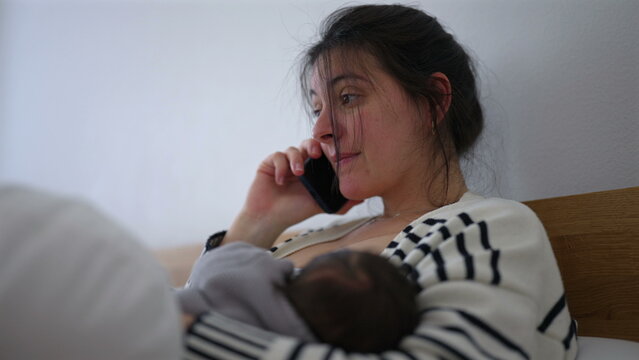Mother multitasking while her baby sleeps peacefully in her arms, speaking on the phone with a thoughtful expression. the quiet balance between caring for a sleeping newborn 