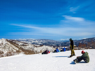 Snowboarders taking a break on a snowy mountain with a vast winter landscape in the background (Togari Onsen, Nagano, Japan)