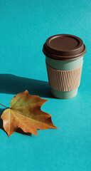 Disposable coffee cup and autumn maple leaf on turquoise background creating a cozy fall mood, perfect for seasonal projects