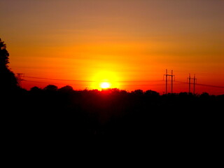 Fiery sunset paints the sky with vibrant orange and yellow over silhouetted trees and power lines