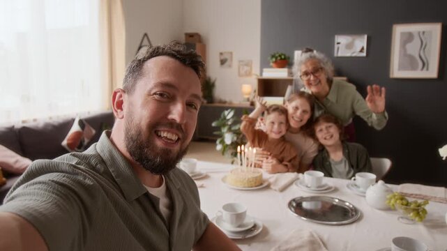 Rack focus POV of smiling man shooting video of extended family consisting of him, wife, two male kids and senior mother-in-law cheerfully waving at camera during birthday celebration at home