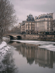 snowy riverbank in paris with haussmann buildings and arched stone bridge under cloudy sky