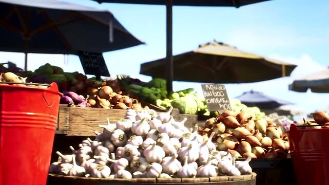 A bustling outdoor market scene with stalls overflowing with fresh produce, featuring large piles of garlic and onions in wooden crates and a bright red bucket in the foreground