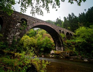 Fototapeta premium Ancient stone arch bridge over a tranquil river. Lush forest surrounds the structure