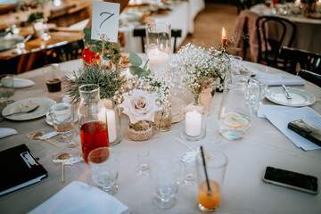 Elegant table setting with candles, flowers, drinks, and a table number in a rustic indoor venue,...