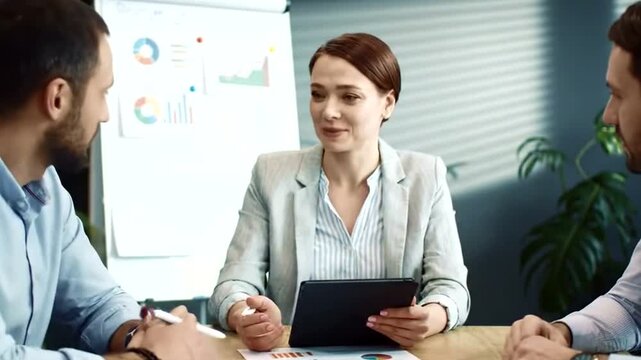 A smiling businesswoman holds a tablet while seated at a table with colleagues during a meeting, discussing charts and graphs