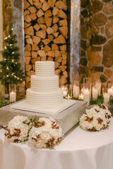 Elegant white wedding cake displayed with floral arrangements near a cozy fireplace during a winter celebration