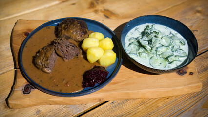 ready to eat -meatballs from chamois meat, potaoes, cranberry jam, brown sauce and cucumber salad, on a blue plate served on a wood board