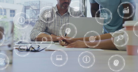 Collaborating team writing and pointing at documents around laptop at meeting table, with icons