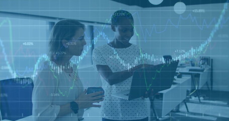 Analyzing charts, two businesswomen discussing data in office, with monitors, laptop and coffee mug
