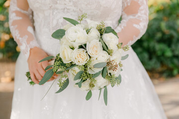 Bridal bouquet arrangement of white roses and greenery held by bride in elegant wedding dress outdoors