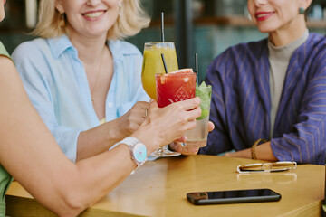 Three middle aged Caucasian women sitting at table clinking colorful cocktails, smiling and engaging in conversation, smartphone and eyeglasses resting on wooden surface