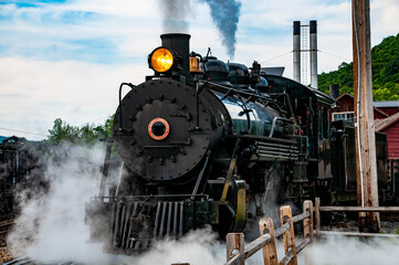 A vintage steam locomotive emits steam as it prepares for departure from a historic railway station. Light clouds contrast with the blue sky, creating a nostalgic atmosphere for onlookers.
