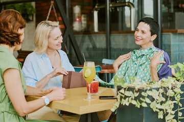 Three middle aged Caucasian women sitting at table in cafe, smiling and talking, one woman gesturing with hands while others listening, drinks and smartphone on table