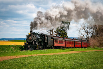 A steam locomotive chugs along a green field, releasing clouds of smoke into the blue sky. The train features wooden passenger cars and is surrounded by rolling hills and trees under bright daylight.
