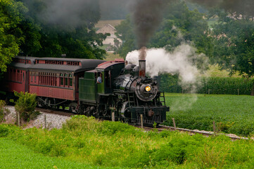 Fototapeta premium A vintage steam train chugs along a scenic railway, billowing smoke against a backdrop of green fields and distant hills. Passengers enjoy the nostalgic ride through beautiful landscapes.