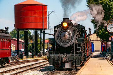 A steam locomotive pulls into the station as passengers eagerly await its arrival. The sun shines brightly, creating a lively atmosphere with people milling about nearby.