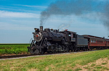 Naklejka premium A vintage steam locomotive moves along a railway track surrounded by lush green fields under a clear blue sky. The train emits black smoke as it journeys through the countryside.