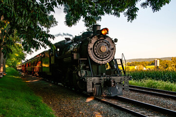 A historic steam locomotive travels along railway tracks surrounded by lush green fields and trees at sunset. The train emits steam and the sun casts a warm glow on the landscape.