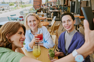 Three Caucasian middle aged women sitting at outdoor cafe table smiling and posing for selfie, holding drinks with straws, enjoying social time together in urban setting