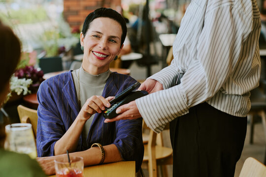 Caucasian middle aged woman smiling while making contactless payment with credit card to young adult Caucasian woman holding payment terminal at outdoor cafe table
