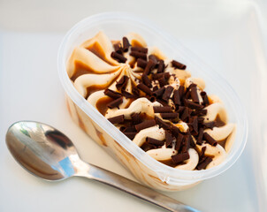There is creamy ice cream with dark caramel and chocolate chips in plastic container. Cold treat, factory-made dessert. Large portion of ice cream packed in disposable container