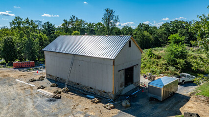 Workers are installing a sleek metal roof atop a newly constructed barn in a tranquil rural setting. The bright sky provides perfect weather for the ongoing project. © Greg Kelton