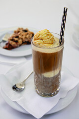 A tall glass of affogato coffee topped with a scoop of vanilla ice cream, served with a striped straw and spoon on a napkin.  Behind; a cherry streuselkuchen, german crumble.