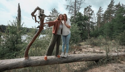 Two smiling women stand together on a fallen tree trunk in a forest near a river, enjoying nature and friendship in a playful, adventurous outdoor moment.