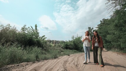 Two women walking on a sandy rural path surrounded by greenery under a bright sky, talking and smiling together while enjoying a peaceful outdoor stroll.